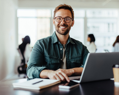 smiling man working on laptop in modern office setting with notebook and coffee cup showcasing productivity tips for seven success strategies