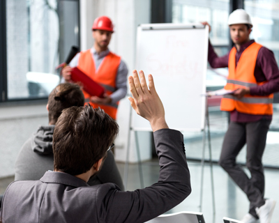 man raising hand during training session with two construction workers in safety vests presenting to a group focusing on safety practices and regulations related to five key areas